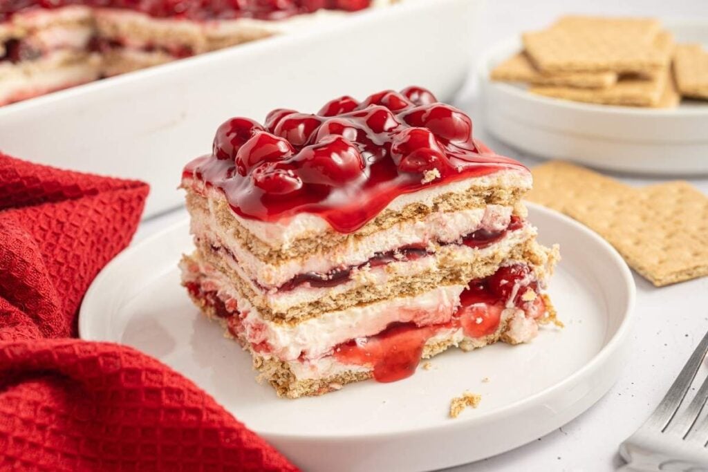 A slice of cherry dessert with layers of graham crackers, cream, and cherry topping on a white plate, with whole graham crackers and a red napkin nearby.