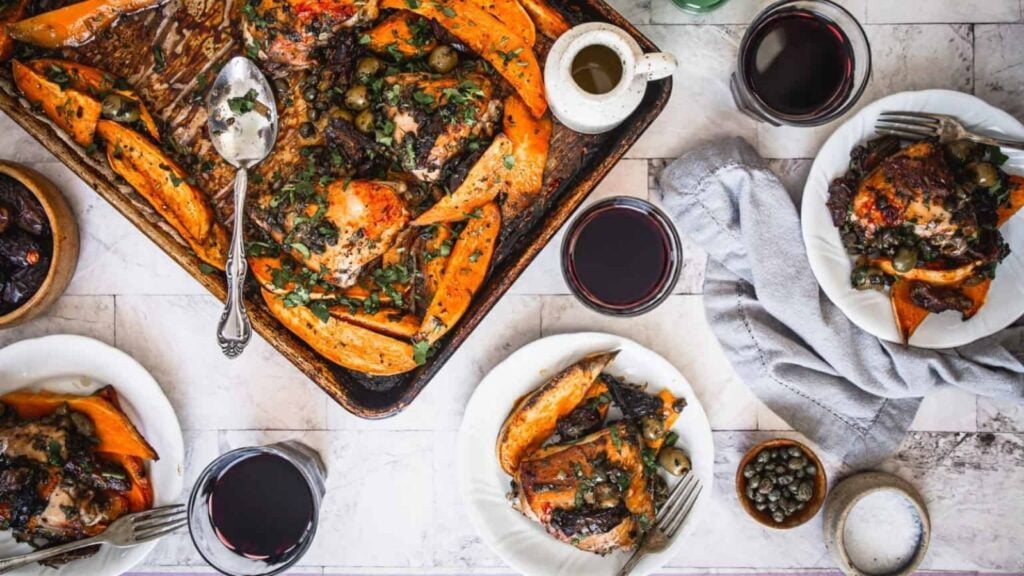 A roasted chicken and sweet potato sheet pan meal is served on plates with glasses of red wine, small bowls of olives and capers, and a gray napkin on a tiled table.