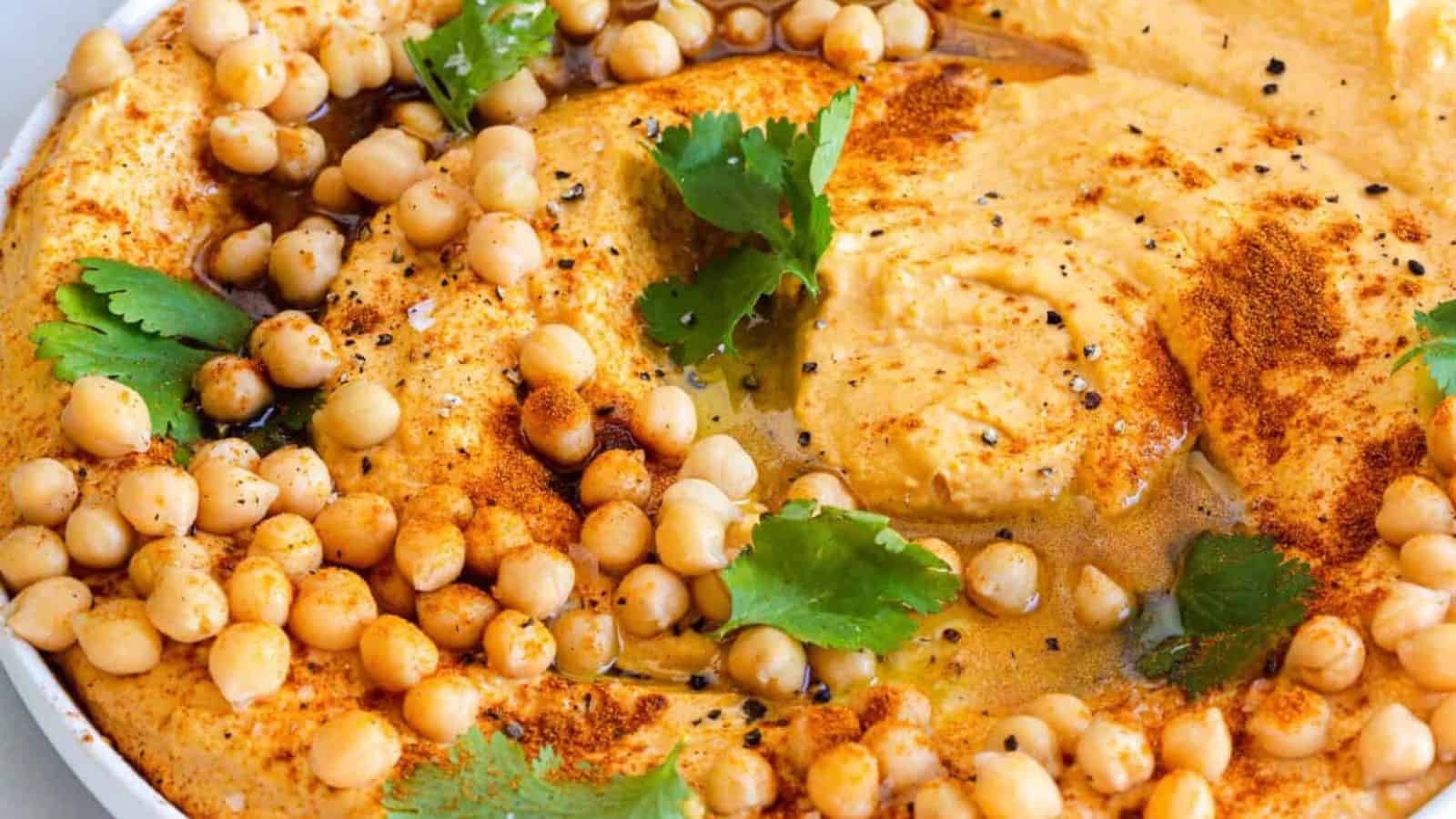 A close-up of a bowl of hummus topped with chickpeas, olive oil, paprika, black pepper, and cilantro leaves. The hummus has a creamy texture and is visually garnished with spices and herbs.