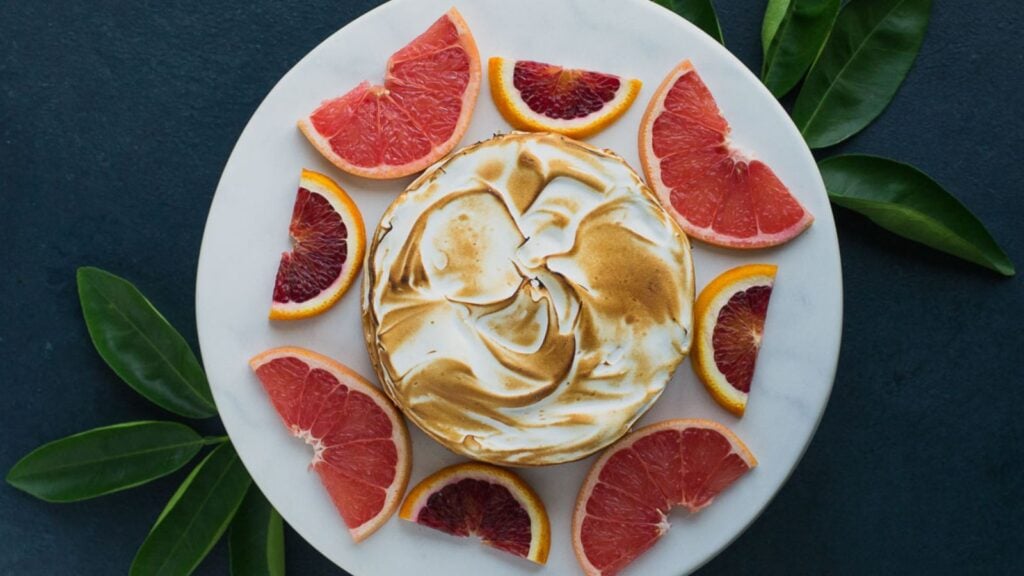 A round dessert with toasted meringue topping is on a white plate, surrounded by grapefruit slices and green leaves, viewed from above.