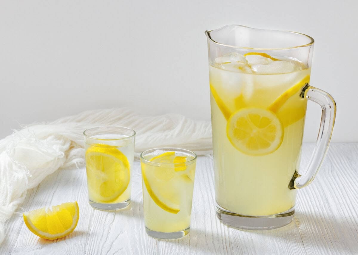 A tall glass pitcher filled with lemonade, ice, and lemon slices stands on a white wooden table, accompanied by two smaller glasses also containing lemonade and lemon. A lemon wedge is placed beside the glasses. A white cloth is draped in the background.