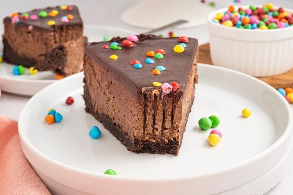 A slice of chocolate cheesecake with a chocolate crust, topped with colorful candy pieces, is served on a white plate. A bowl of candies is in the background.