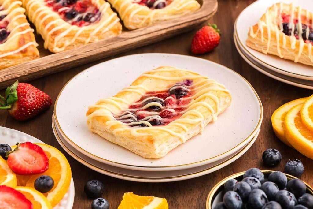 A rectangular fruit danish pastry with icing drizzle on a white plate, surrounded by fresh strawberries, blueberries, and orange slices on a wooden table.