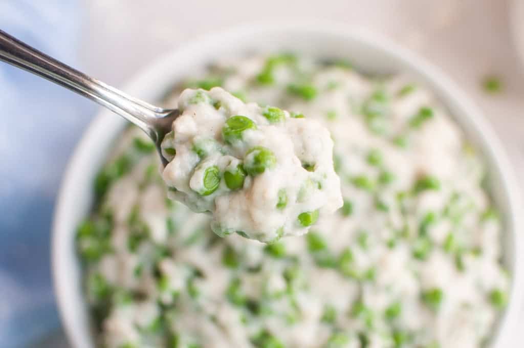 A spoonful of creamy green peas and rice is held above a white bowl filled with the same dish.