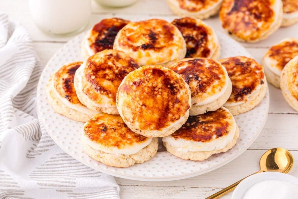 A plate of round, browned Welsh cakes is stacked on a white polka dot plate, set on a table with a striped napkin and a gold spoon nearby.