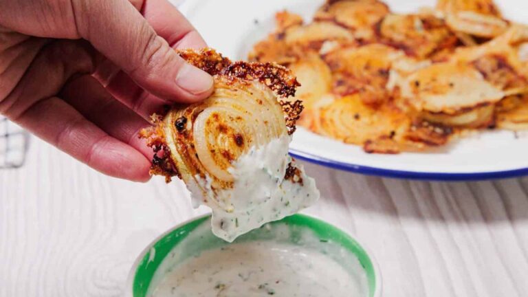A hand dips a crispy, baked onion slice into a bowl of creamy dipping sauce, with more onion slices on a plate in the background.