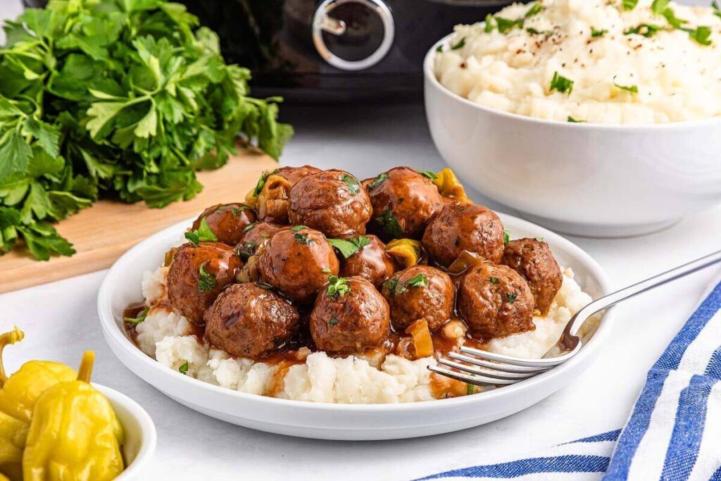 A plate of meatballs in brown gravy served over mashed potatoes, garnished with herbs, with a fork and a bowl of mashed potatoes in the background.