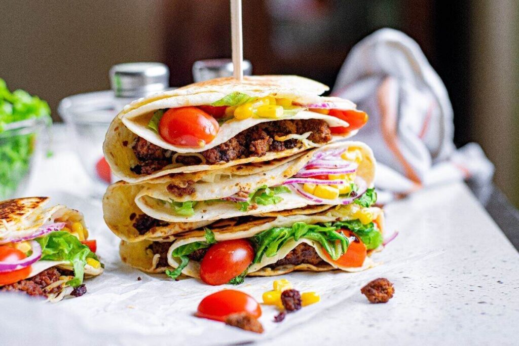 Three folded tortillas filled with ground beef, lettuce, tomatoes, red onions, and cheese are stacked on a countertop, with a skewer holding them together.