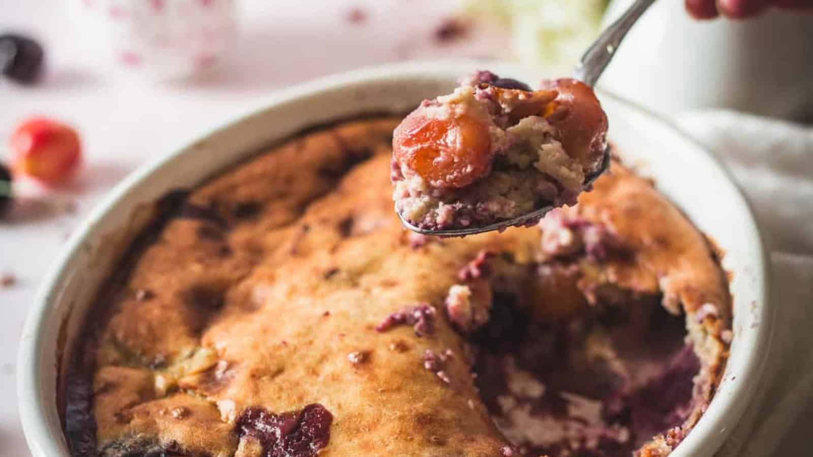 A baked fruit cobbler in a round white dish with a portion already served. A spoon holds up a serving, showing fruit filling and golden-brown crust. Cherries and crumbs are visible on the table in the background.