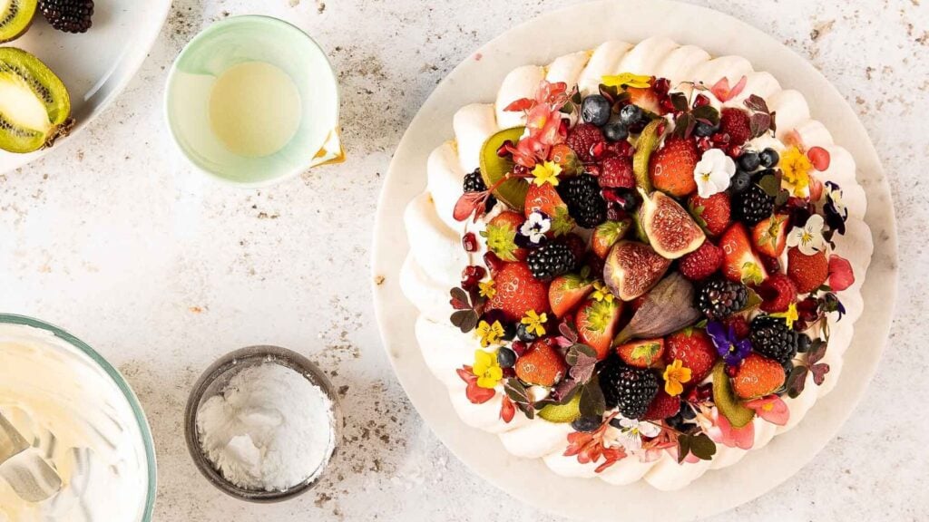 A pavlova dessert topped with assorted fresh berries, figs, kiwi slices, and edible flowers sits on a white plate surrounded by bowls of ingredients.