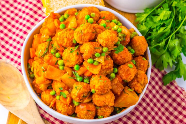 A bowl of cooked vegetable curry with peas and round vegetarian meatballs, garnished with herbs, sits on a red checkered cloth next to fresh parsley and a wooden spoon.