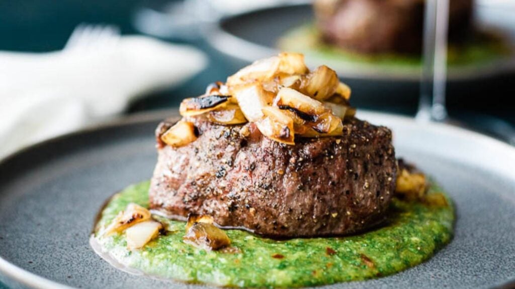 A grilled steak topped with sautéed onions sits on a bed of green sauce on a dark plate.