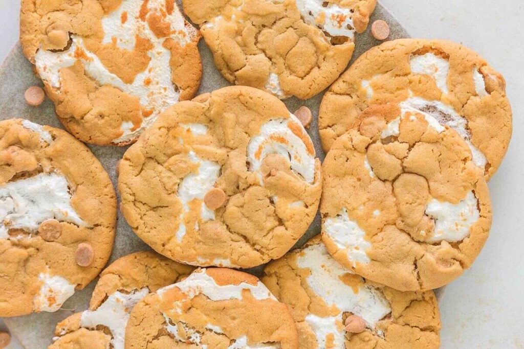 A close-up of several peanut butter cookies with swirls of melted marshmallow and butterscotch chips on a white surface.