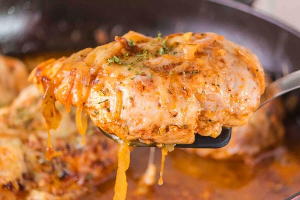 A spatula holds a succulent piece of French onion chicken, smothered in cheese and herbs, with sauce dripping tantalizingly.