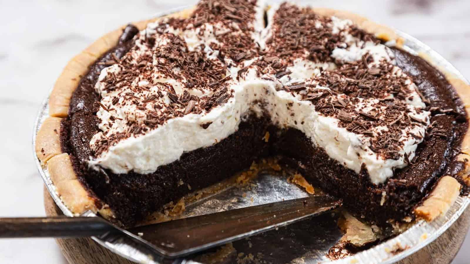A chocolate cream pie in a metal pie tin, topped with whipped cream and chocolate shavings. A slice has been cut and a metal pie server is visible under the empty space.