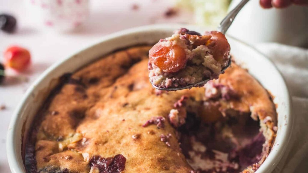 A close-up of a baked cherry cobbler in a white dish with a spoon lifting out a portion containing cherries and crust.