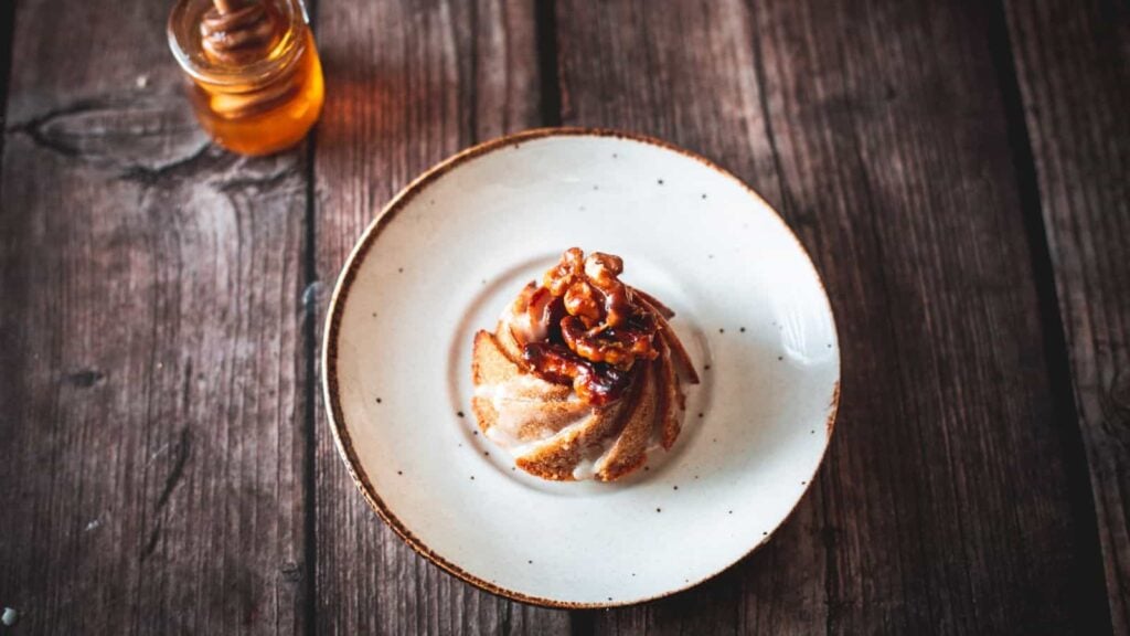 A small bundt cake topped with walnuts and syrup sits on a white plate, with a jar of honey in the background on a wooden table.