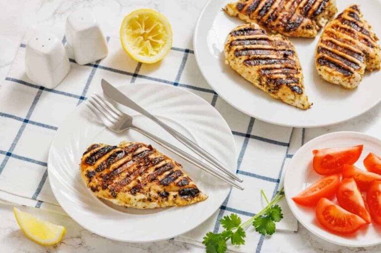 Grilled chicken breasts on white plates, sliced tomatoes, a halved lemon, cilantro, salt and pepper shakers, and cutlery on a checkered cloth.
