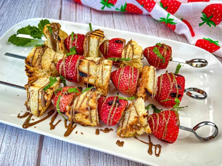 Skewers of grilled bread and fresh strawberries, drizzled with chocolate, and garnished with mint, arranged on a white rectangular platter, with a strawberry patterned napkin visible in the background.