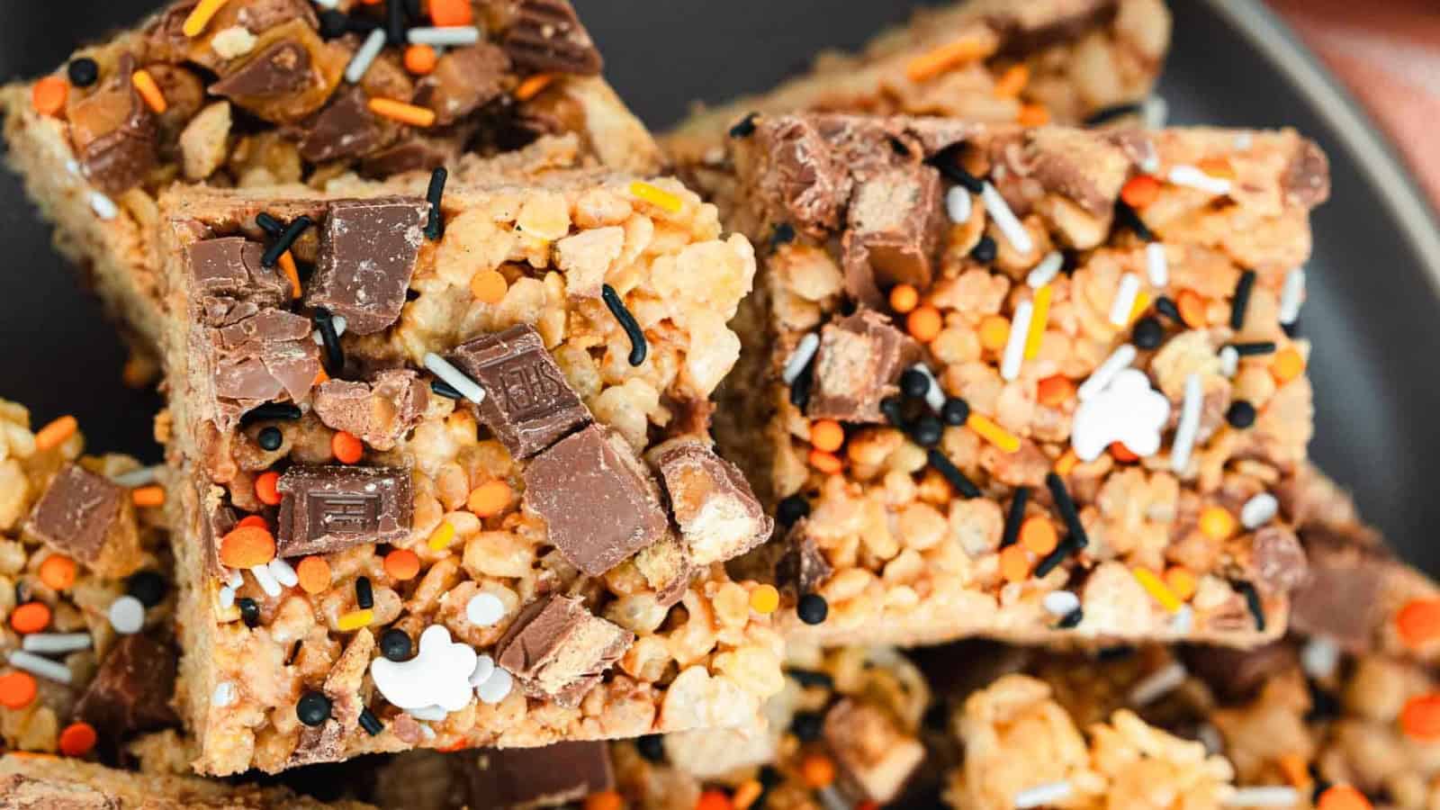Rice crispy treats topped with chocolate pieces and colorful Halloween-themed sprinkles on a dark plate.