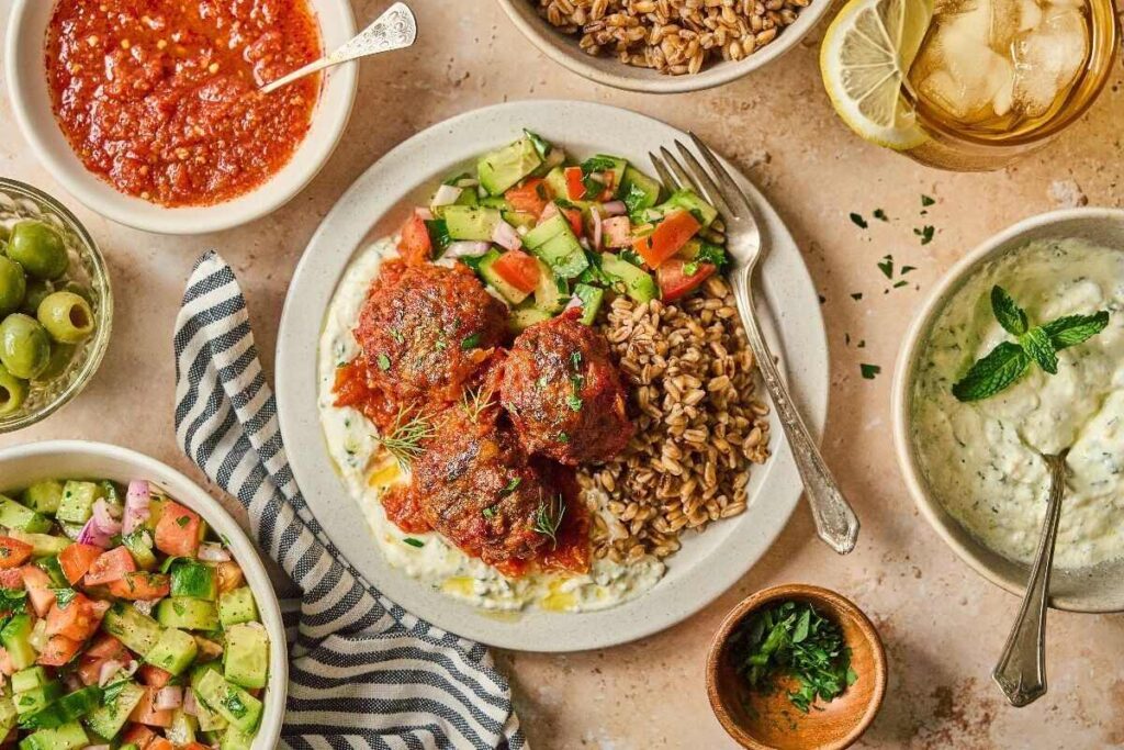 A plate with meatballs in tomato sauce, farro, cucumber-tomato salad, and yogurt sauce, surrounded by bowls of salad, sauce, olives, and a glass of iced drink.