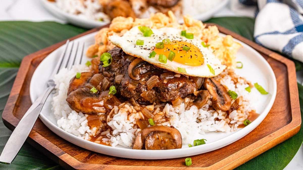 A hearty breakfast plate of rice topped with a hamburger patty, brown gravy with onions and mushrooms, a fried egg, and chopped green onions, served with a fork on the side.