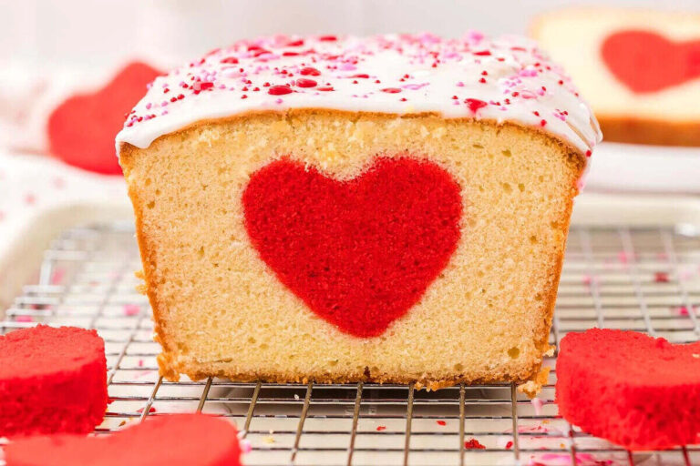 A loaf cake with white icing and pink sprinkles features a red heart shape in the center, displayed on a cooling rack with red heart-shaped cake pieces around it.