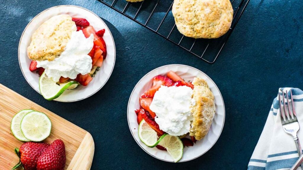 Two plates of strawberry shortcake with whipped cream and lime slices sit on a dark surface, with extra biscuits on a cooling rack and fresh strawberries and limes on a cutting board nearby.