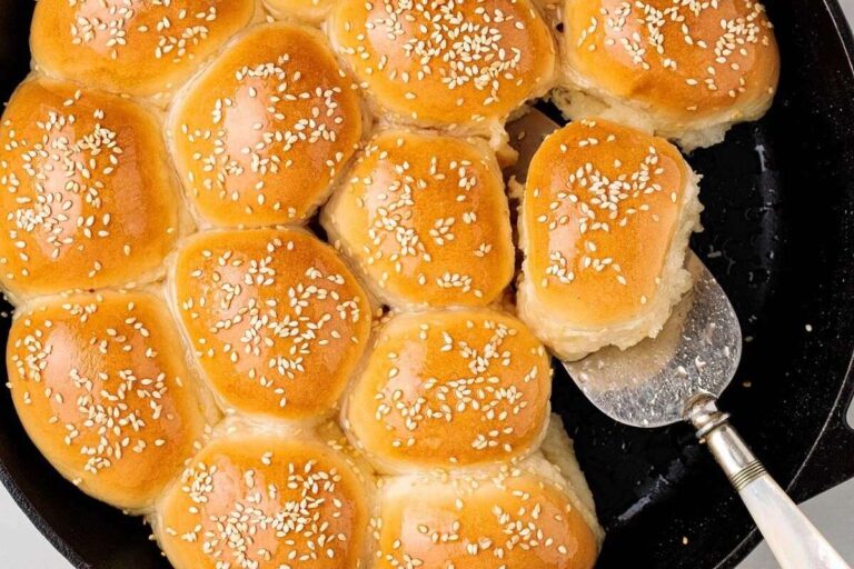 A batch of golden brown, sesame-topped dinner rolls in a pan, with one roll being lifted out using a metal spatula.