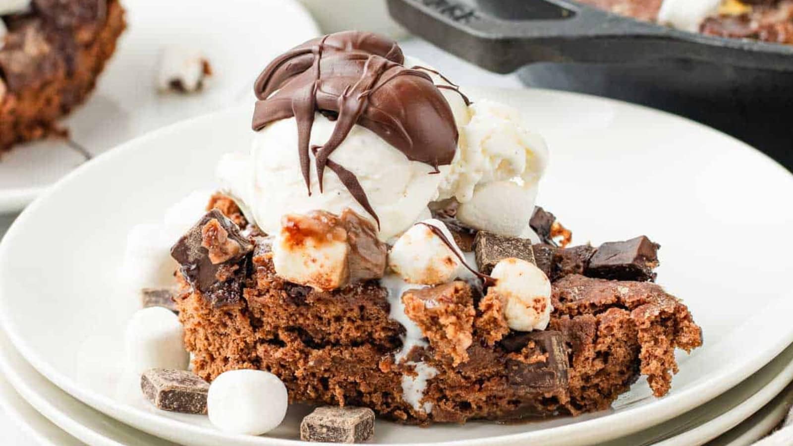 A chocolate brownie topped with ice cream, marshmallows, and chocolate drizzle on a white plate.