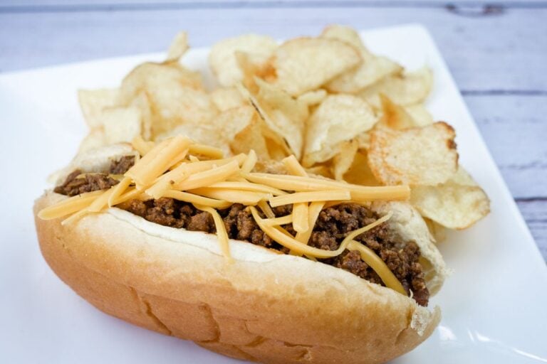 A chili cheese hot dog topped with shredded cheddar cheese is served on a plate with a side of potato chips.
