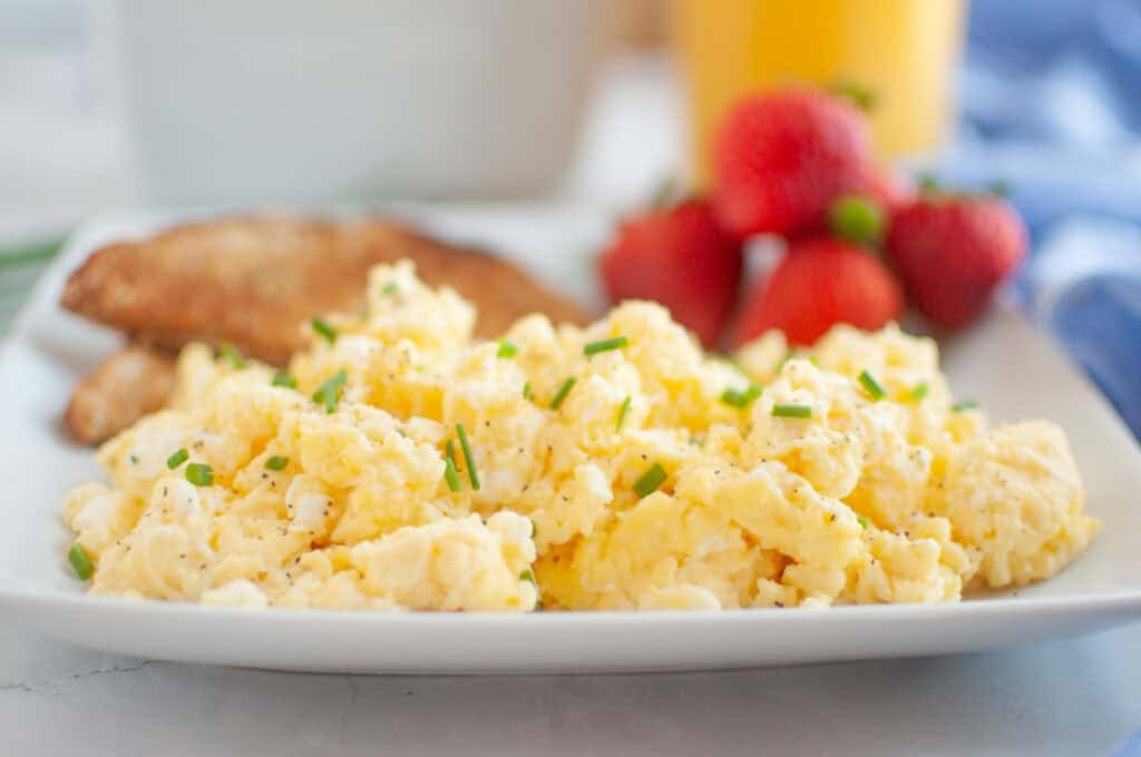 A plate of scrambled eggs garnished with chives, served with toast and strawberries, with a glass of orange juice in the background.
