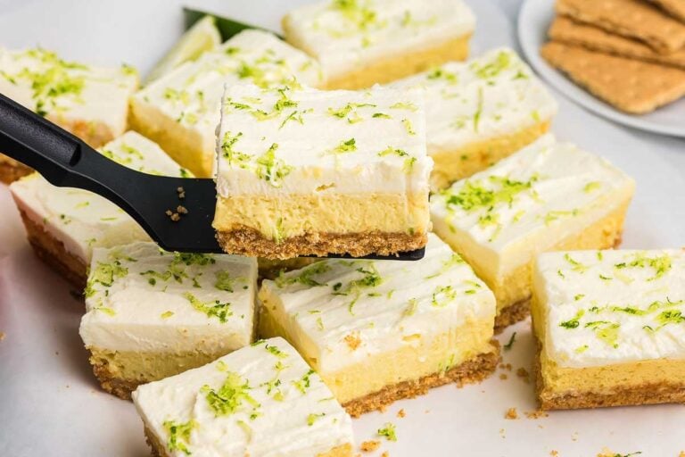 A spatula lifts a square piece of key lime cheesecake bar topped with whipped cream and lime zest from a group of similar bars on a white surface.