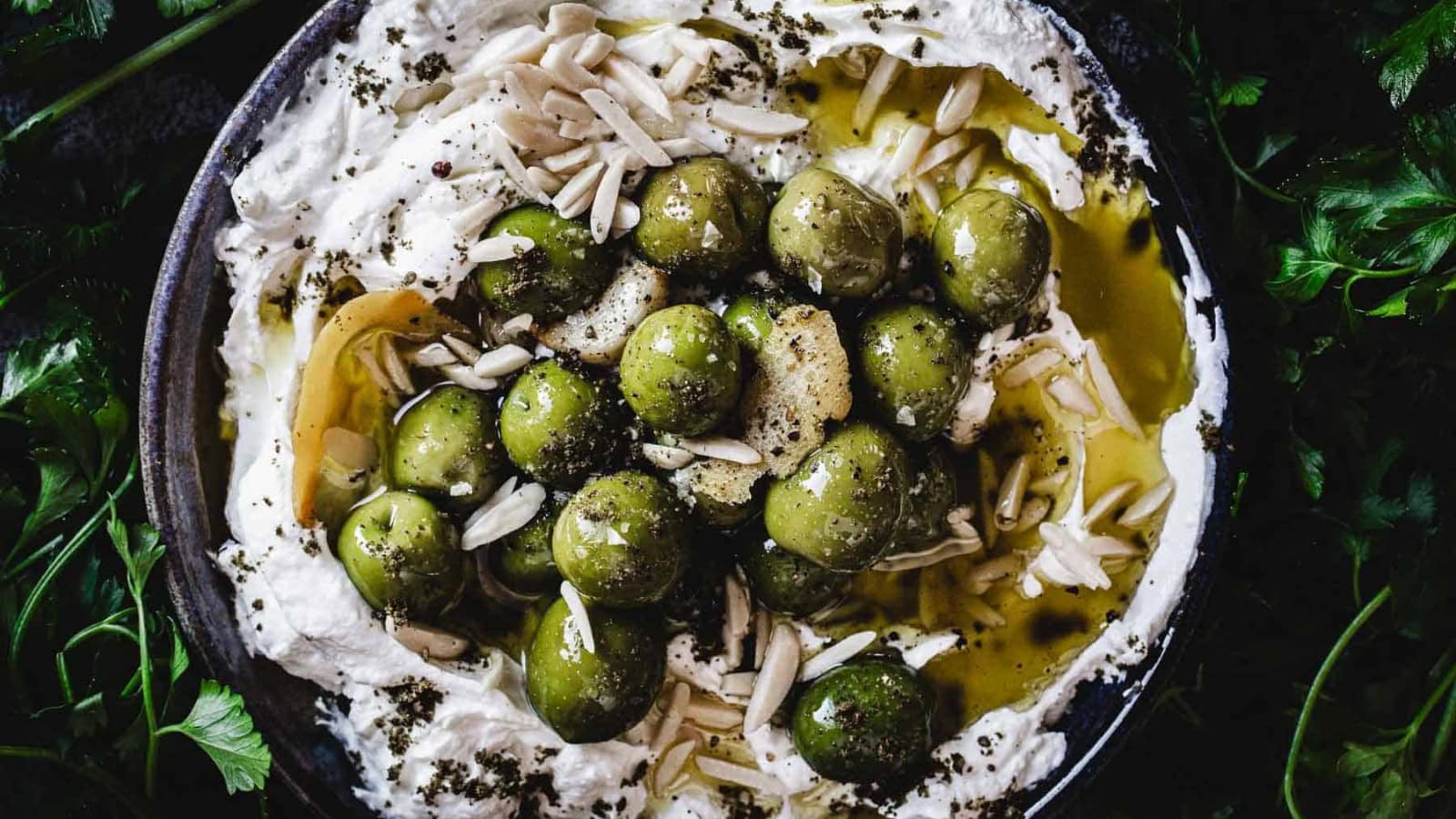 A bowl of whipped labneh topped with green olives, olive oil, slivered almonds, and herbs, surrounded by fresh parsley leaves.