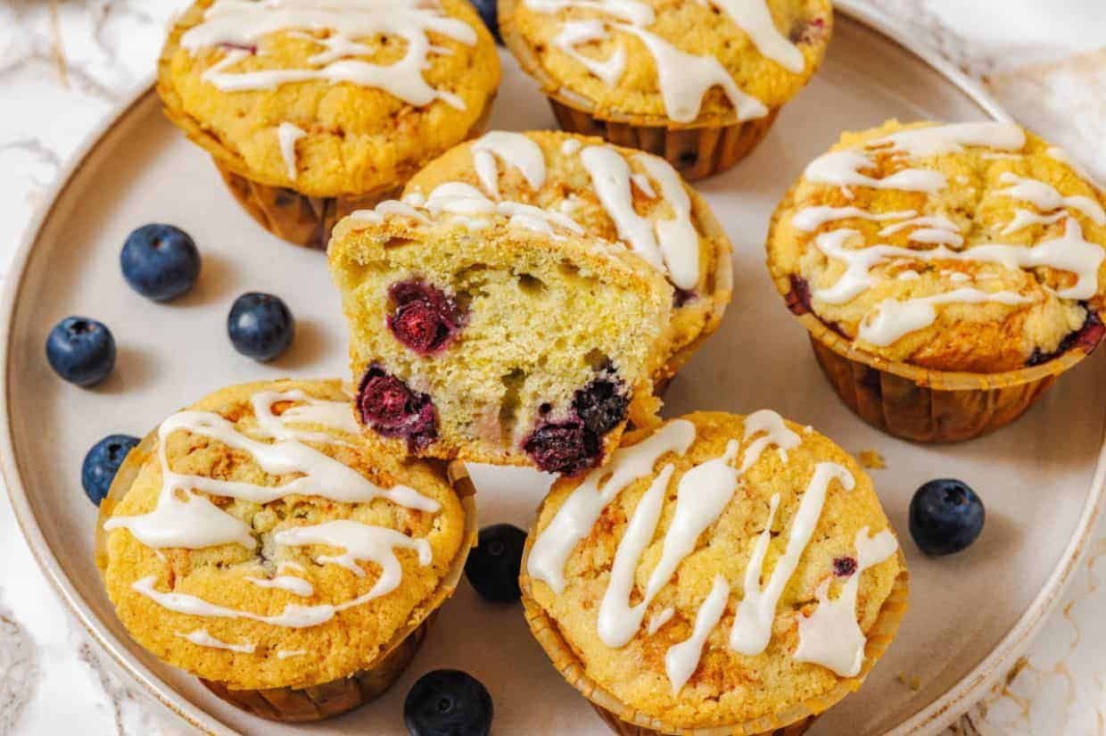 A plate with five Lemon Blueberry Muffins, one muffin cut in half showing the blueberries inside, and some loose blueberries around them.