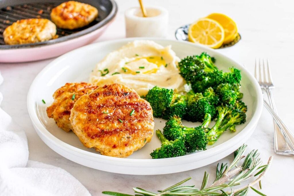 Plate with two grilled lemon herb chicken patties, a serving of mashed potatoes topped with butter, and steamed broccoli.
