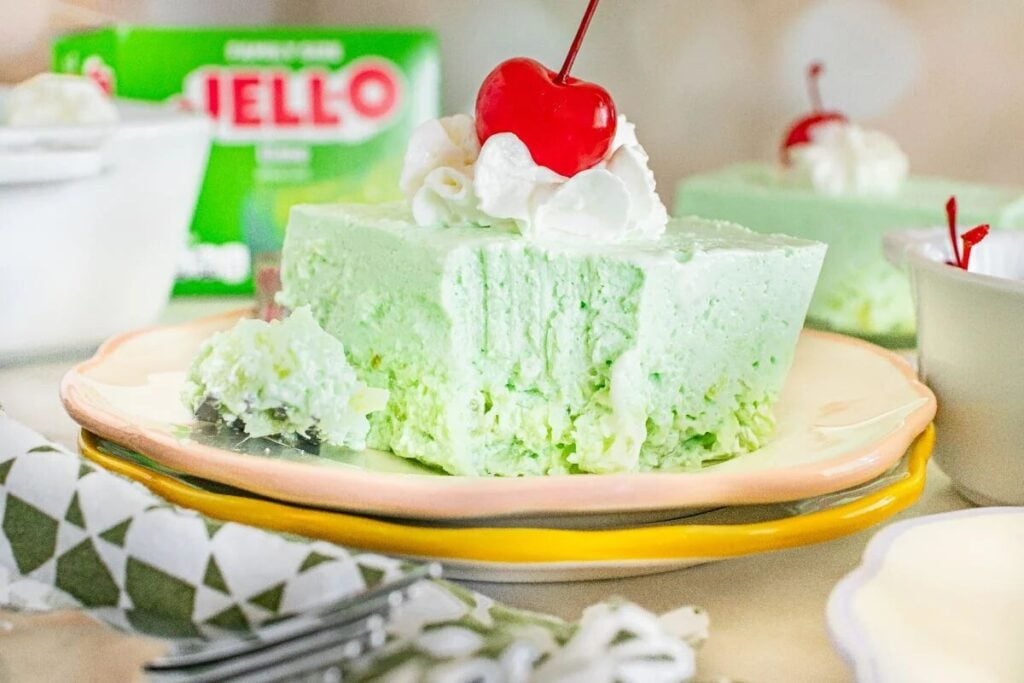 A slice of light green Jello dessert topped with whipped cream and a cherry sits on a plate, with a bite taken out and a Jello box in the background.