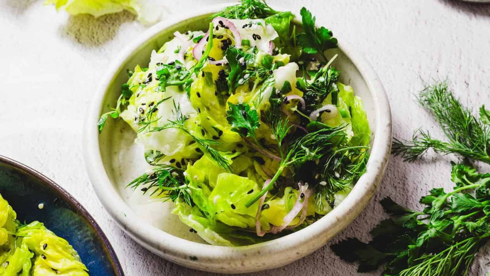A bowl of salad containing leafy greens, fresh herbs, sliced onions, and black sesame seeds on a light surface. Another dish with similar contents is partially visible to the side. There are scattered herbs around the bowl.