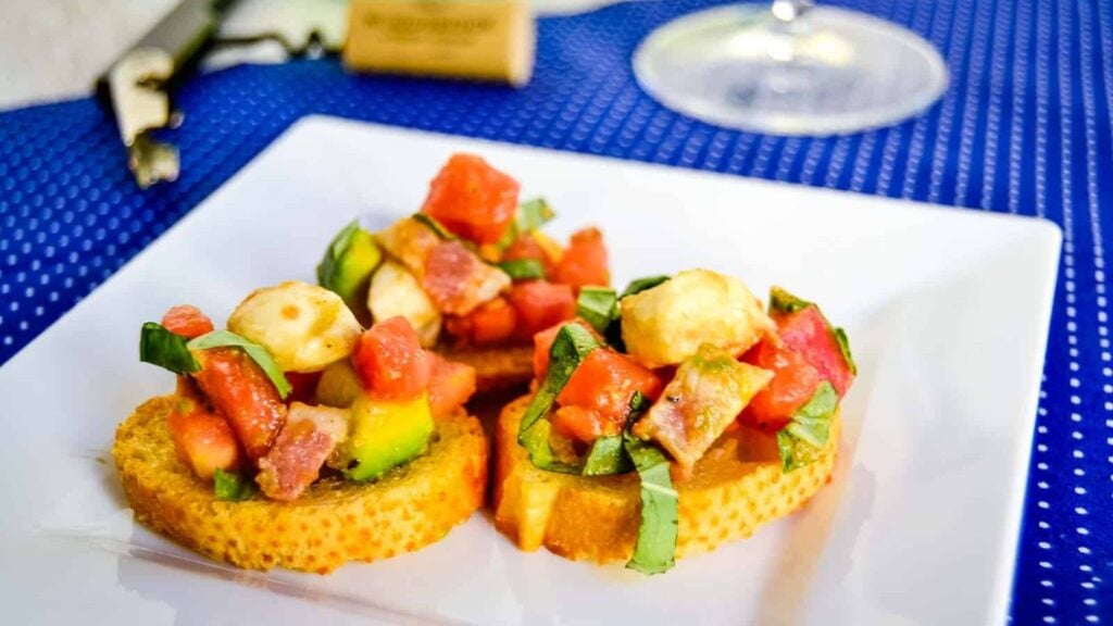 Two pieces of toasted bread topped with diced tomatoes, mozzarella, avocado, and basil on a white plate, placed on a blue polka dot surface.