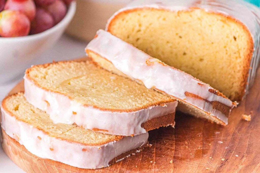 Sliced glazed pound cake on a wooden cutting board with a bowl of grapes in the background.