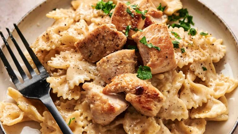 A plate of bowtie pasta with creamy sauce, topped with grilled chicken pieces and garnished with chopped parsley, with a black fork on the side.