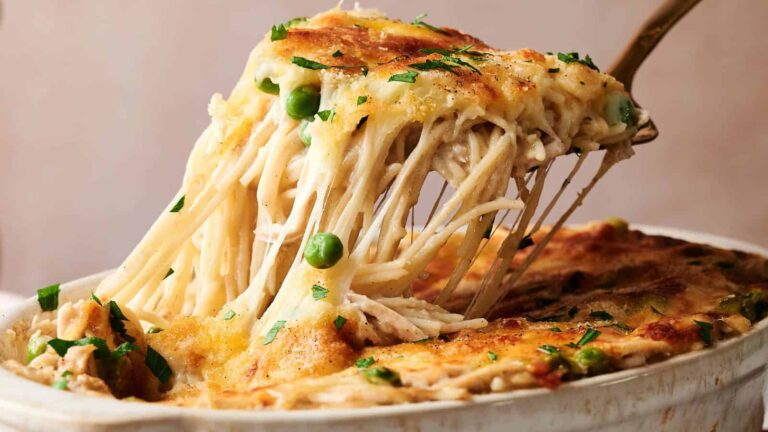 A serving of cheesy baked pasta with green peas is being lifted from a casserole dish, showing melted cheese and a golden-brown top.
