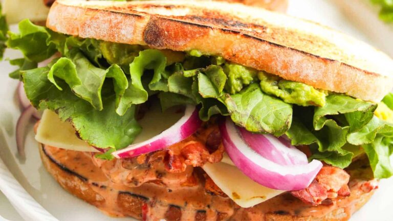 A close-up of a sandwich with toasted bread, leafy lettuce, sliced red onions, cheese, bacon, creamy sauce, and mashed avocado.