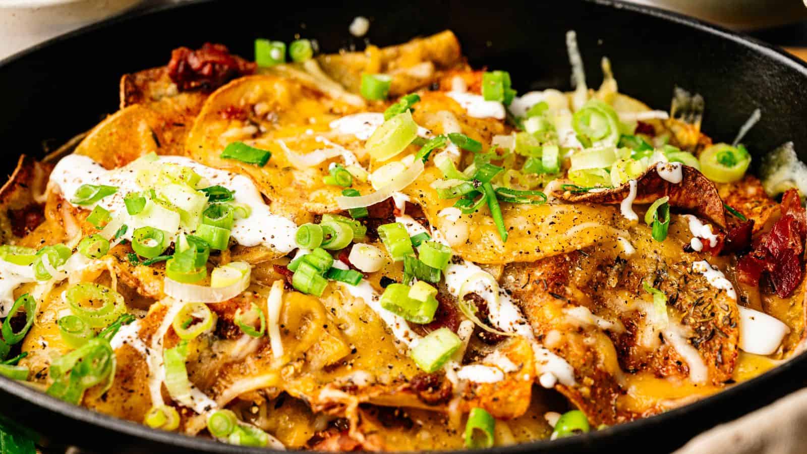A close-up of a skillet filled with cheesy nachos topped with sliced green onions, melted cheese, sour cream, and various seasonings.
