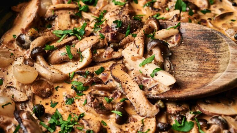 A close-up of creamy mushroom stroganoff garnished with chopped parsley, with a wooden spoon resting in the dish.