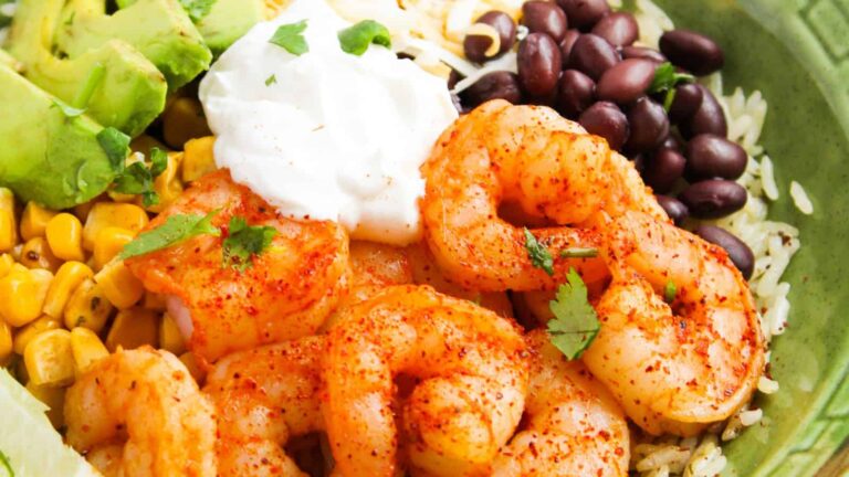 A close-up of a shrimp bowl with seasoned shrimp, black beans, corn, avocado, shredded cheese, rice, and a dollop of sour cream, garnished with cilantro.