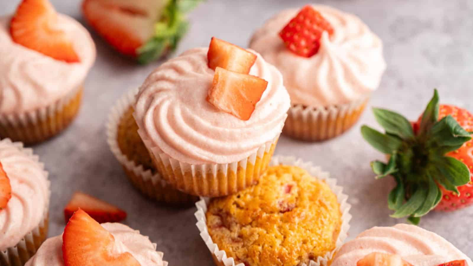 Several cupcakes topped with pink strawberry frosting and fresh strawberry pieces are arranged on a light surface, with whole and sliced strawberries nearby.