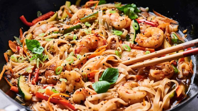 A bowl of shrimp stir-fry with rice noodles, vegetables, and sesame seeds, garnished with green onions and basil, with chopsticks resting on the side.