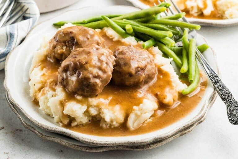 A plate with mashed potatoes topped with brown gravy and meatballs, served with green beans and a fork.