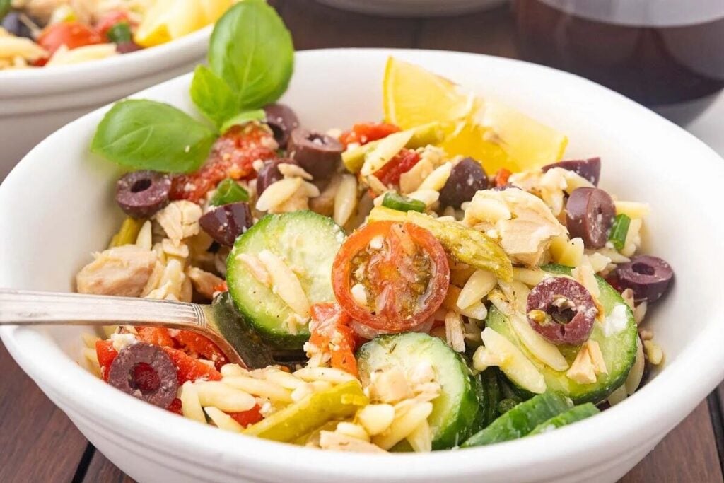 A bowl of orzo pasta salad with sliced cucumbers, tomatoes, kalamata olives, basil leaves, and a lemon wedge, served with a fork.